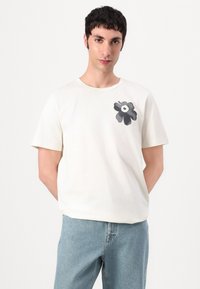 Tricou din bumbac de culoare crem, cu mâneci scurte, având un mare design floral negru în zona stângă a pieptului. Guler clasic rotund.