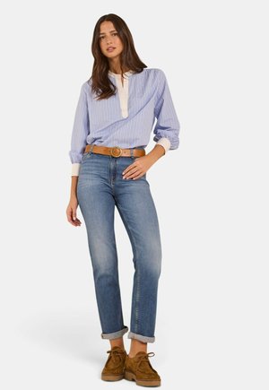 Femme debout, main sur la hanche, portant un chemisier rayé bleu, un jean retroussé, une ceinture marron et des chaussures en daim marron sur fond blanc.
