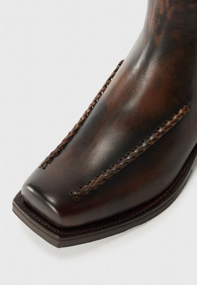 Bota de cuero marrón con punta cuadrada, con detalle trenzado a lo largo de los lados y una superficie texturada. Diseño de suela sólida y resistente.