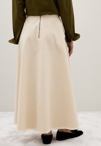 Femme portant une longue jupe beige avec une fermeture éclair au dos, un haut vert olive et des escarpins plats noirs, se tenant sur un sol recouvert de moquette blanche.