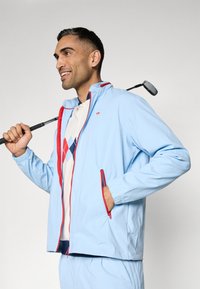 Chaqueta azul claro con acentos rojos, bolsillos con cremallera y puños elásticos. Lleva encima un polo crema con un patrón de diamantes en azul y rojo.
