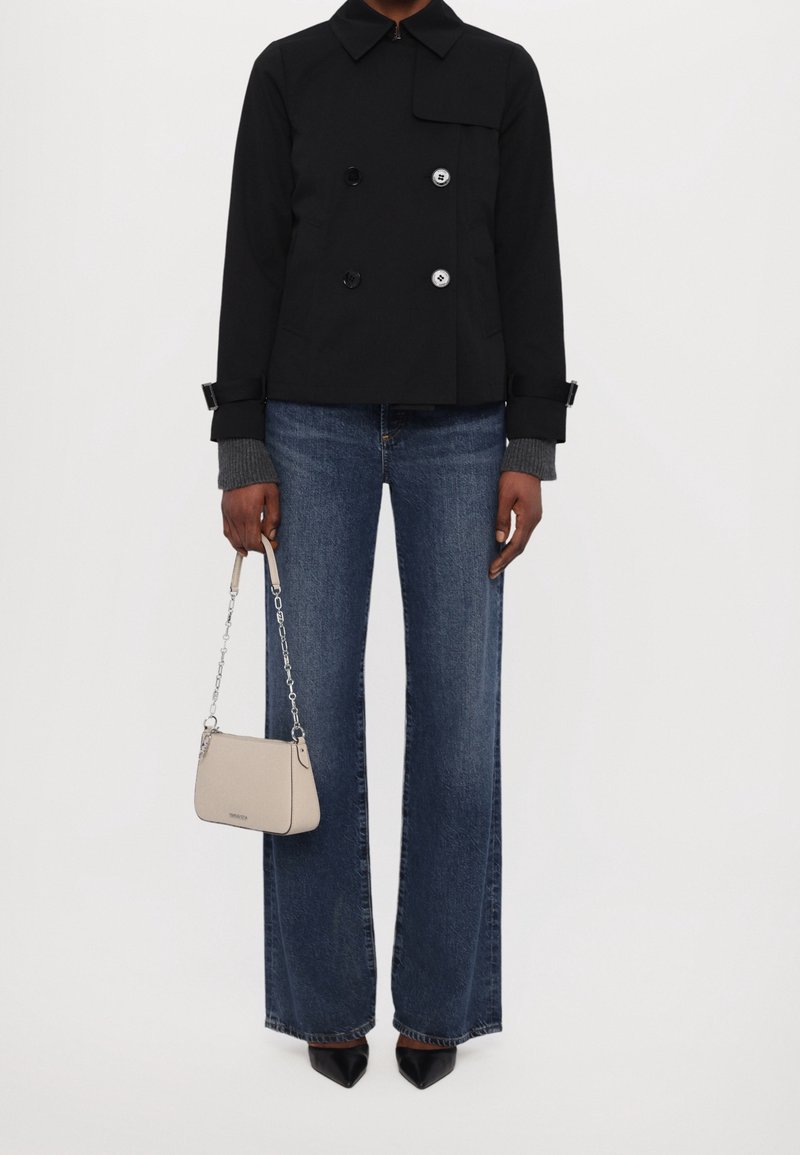 Personne portant une veste noire croisée, un jean bleu à jambes larges, des chaussures pointues noires, et tenant un petit sac à main beige avec une bandoulière chaîne.