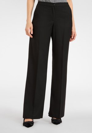 Pantalons noirs à jambes larges avec une texture lisse, une fermeture à bouton sur le devant et une coupe ajustée, associés à des chaussures plates noires.