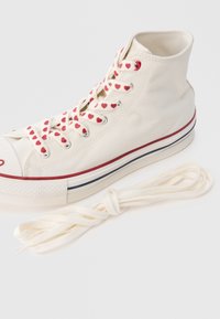 Λευκό καμβαδένιο ψηλό αθλητικό παπούτσι με κορδόνια σε κόκκινο χρώμα με σχέδιο κόκκινων καρδιών, μεσαία σόλα με κόκκινες και μπλε ρίγες και λαστιχένια μύτη. Περιλαμβάνει επιπλέον λευκά κορδόνια.