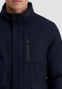 Marineblauwe zip-up jack met hoge kraag, voorzien van een ritszak op de borst en twee voorzakken. gemaakt van een gestructureerde, duurzame stof.