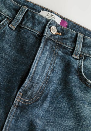 Jeans typu flare w ciemnoniebieskim dżinsie z subtelnym przecieraniem, kontrastującymi pomarańczowymi szwami i srebrnym zapięciem na guzik. Metka widoczna wewnątrz.