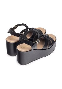 Sandales à plateforme noires avec de larges brides en cuir croisées et une boucle en ton or. Semelle en liège de couleur beige, avec un talon surélevé.