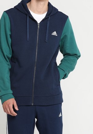 Man draagt een marineblauwe Adidas zip-up hoodie met groene mouwen en bijpassende marineblauwe Adidas trainingsbroek.