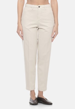 Donna che indossa pantaloni beige a gamba dritta con passanti per cintura e scarpe marroni senza lacci, in piedi contro uno sfondo chiaro e semplice.