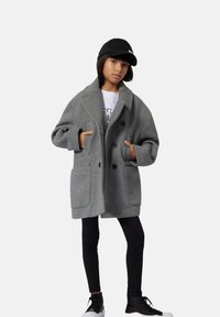 Szary płaszcz z mieszanki wełny o podwójnym zapięciu, z dużymi kieszeniami i klapami, noszony na czarne legginsy i sneakersy.