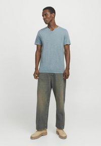 Lyseblå, kortærmet, tekstureret T-shirt med V-hals og to knapper, parret med løse, slidte grønne jeans og beige støvler.
