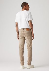 Jenas de ganga bege com um corte regular, pernas afuniladas e um crachá de couro castanho na parte de trás. Combinados com uma t-shirt branca lisa e sapatos brancos.