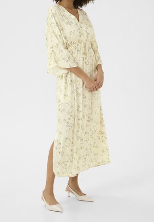 Kvinde iført en lys gul blomsterprint midi-kjole med brede ærmer og snørret talje, stylet med beige lave hælede slingback-sko.