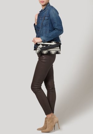 Femme portant une veste en jean bleu, pantalon en cuir marron foncé, bottines beiges en daim à talons hauts, tenant un sac à bandoulière noir et blanc à motifs.