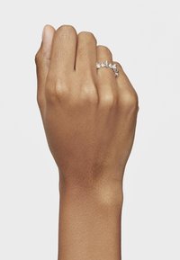 Srebrny pierścionek z wieloma małymi, przejrzystymi kamieniami, o falistym wzorze, noszony na dłoni o średnim brązowym odcieniu skóry.