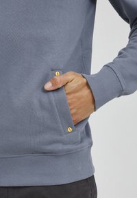 Felpa grigia con polsini e orlo a coste. Presenta una tasca laterale con dettaglio di bottone a scatto giallo. Tessuto liscio e vestibilità regolare.