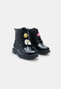 Botas de tobillo de charol negro con puntera redonda, cordones en la parte delantera y charms decorativos coloridos: cara sonriente, flor y acentos de arcoíris.