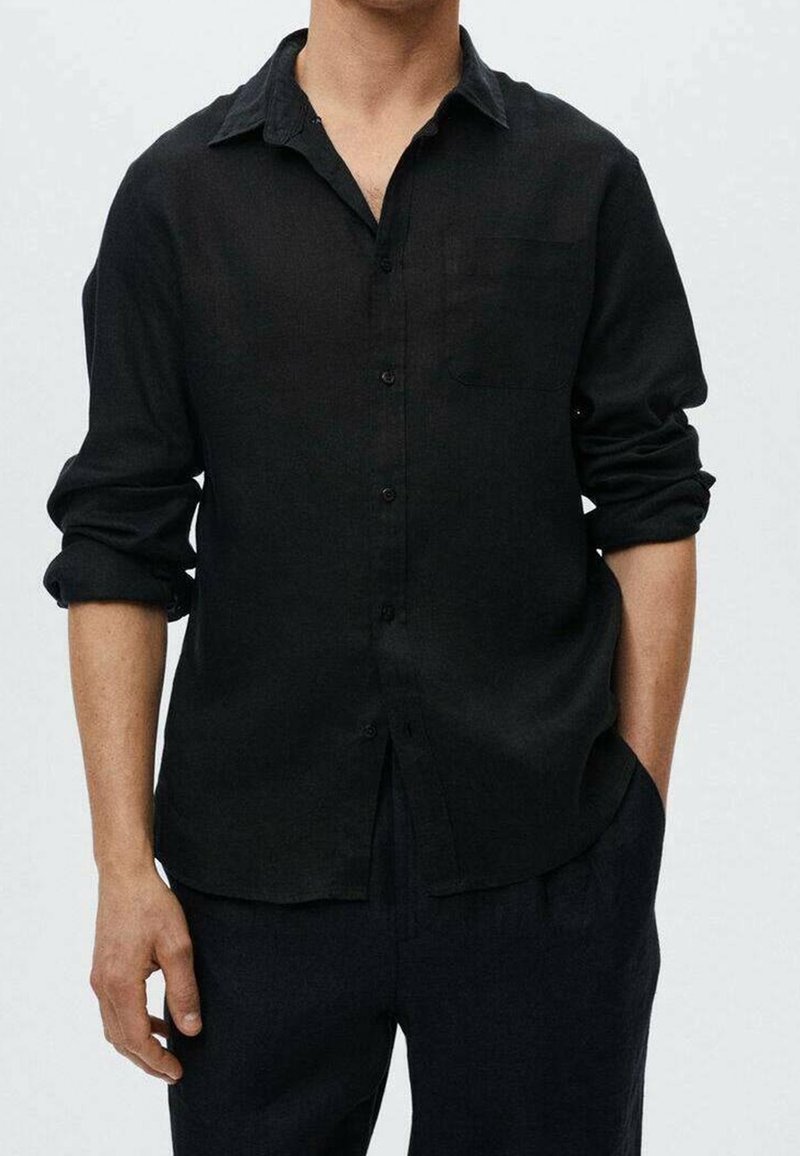 Camisa de lino negra con cuello, frente abotonado, mangas largas remangadas y un solo bolsillo en el pecho, combinada con pantalones oscuros.