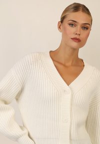 Cardigan bianco lavorato a maglia con scollatura a V, caratterizzato da un motivo a coste testurizzato e due bottoni sul davanti. Maniche lunghe e larghe conferiscono volume.