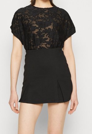 Kvinde iført en sort transparent bluse med blomsterblonde sammen med en sort højtaljet mininederdel med forreste læg.