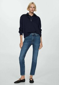 Pulover bleumarin cu guler în formă de jumătate de fermoar, asortat cu blugi strâmți și pantofi negri fără toc. Îmbrăcămintea are un design și texturi minimaliste.