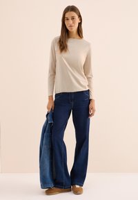Beige long-sleeve top paired with dark blue wide-leg jeans, holding a denim jacket. Outfit features simple lines and minimalistic design.