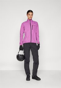 Giacca impermeabile viola con zipper frontale, guanti neri, pantaloni neri con accenti laterali e un casco nero tenuto in mano.