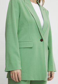 Grön blazer i slätt tyg, med en enda brun knapp, slagkragar och sidofickor, med en avslappnad passform och långa ärmar.