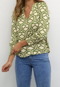 Kvinde justerer ærmet på en grøn bluse med geometrisk mønster, der er parret med blå jeans foran en simpel hvid baggrund.