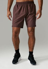 Brune atletiske shorts med elastisk talje og lommer. Glat tekstur, knæ-længde, parret med sorte sko og sokker.