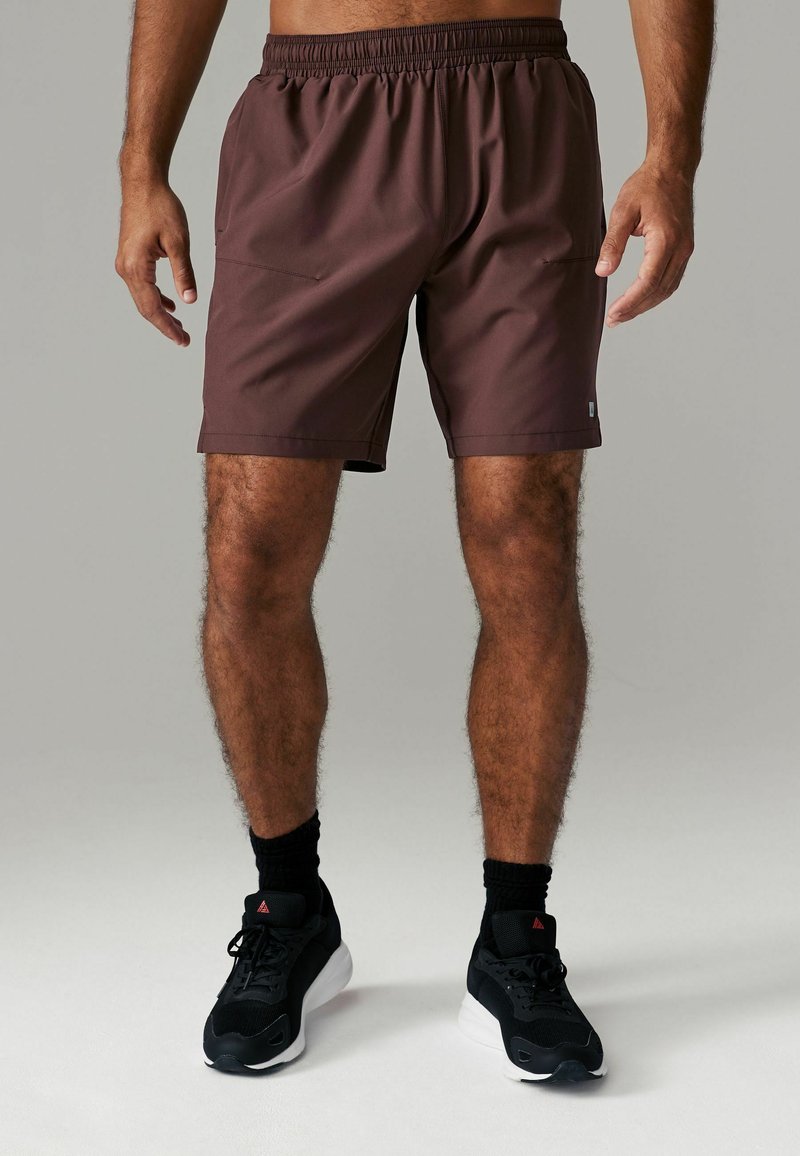 Brune atletiske shorts med elastisk talje og lommer. Glat tekstur, knæ-længde, parret med sorte sko og sokker.
