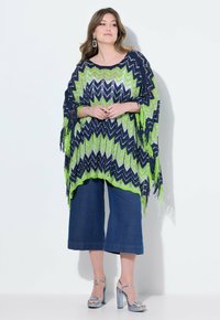 Stickad poncho i marinblått, grönt och vitt zigzag-mönster, med fransade kanter. Matchad med vida, avklippta jeans och silverfärgade klackar.