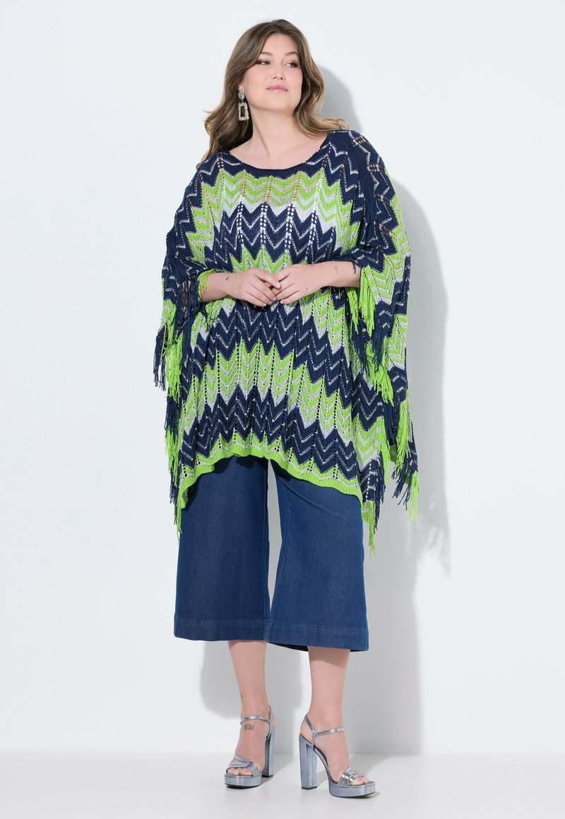 Stickad poncho i marinblått, grönt och vitt zigzag-mönster, med fransade kanter. Matchad med vida, avklippta jeans och silverfärgade klackar.