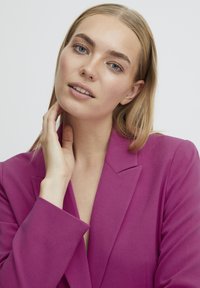 Fuksija sako s oštrim reverima, teksturiranom tkaninom i dugim rukavima. Model dodiruje vrat, pokazujući čist, minimalistički stil.