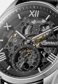 Skeleton horloge met een zwart en zilver metalen ontwerp. Bevat Romeinse cijfers, ingewikkelde tandwieldetails en een gouden accent met leren band.
