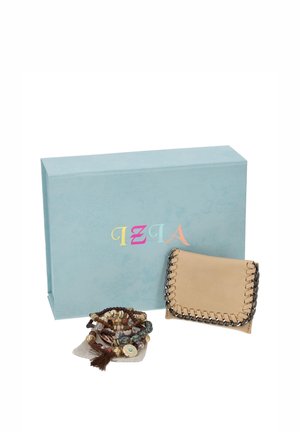 Una cartera beige con solapa y adornos de cadena, junto a una pila de pulseras de cuentas, está delante de una caja de regalo azul claro.