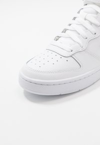 Vit sportskor med perforerat tåhuvud, slät läderoversidan, tjock sula och platta snören, framställda mot en enkel bakgrund.