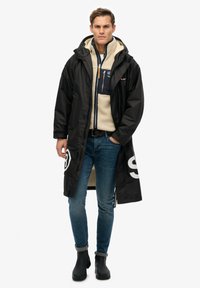 Abrigo largo negro impermeable con capucha, superpuesto a una chaqueta de forro polar beige. Presenta unos vaqueros de mezclilla y botas negras hasta el tobillo. Con grandes acentos gráficos.