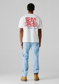 Λευκό βαμβακερό μπλουζάκι με κόκκινο και μπλε κείμενο στην πλάτη που γράφει "RED BULL RACING SPEED AND PRECISION", συνδυασμένο με ανοιχτόχρωμο τζιν και καφέ μπότες.