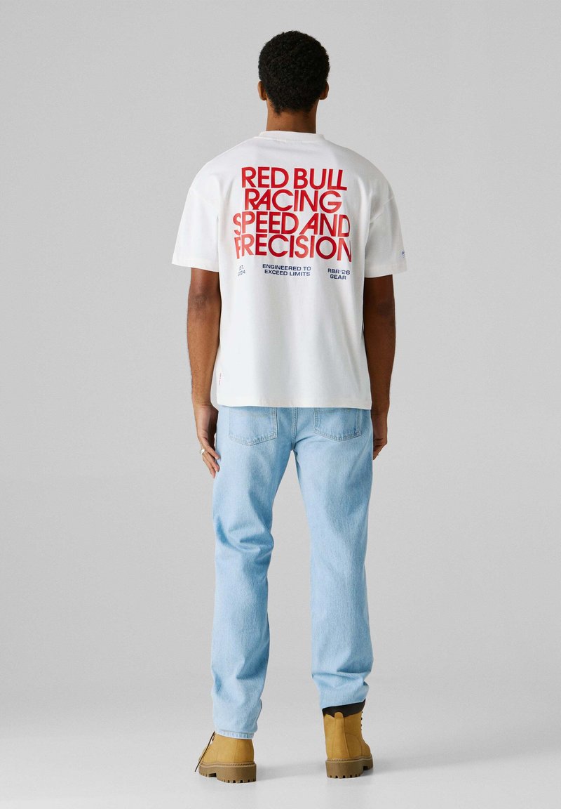 Λευκό βαμβακερό μπλουζάκι με κόκκινο και μπλε κείμενο στην πλάτη που γράφει "RED BULL RACING SPEED AND PRECISION", συνδυασμένο με ανοιχτόχρωμο τζιν και καφέ μπότες.