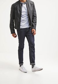 Giacca di pelle nera con dettagli trapuntati sulle spalle, t-shirt grigia, jeans blu scuro e sneakers bianche con calzini grigi.