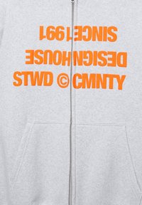 Svijetlosiva dukserica na patent s jarkim narančastim tekstom "STWD © CMNTY DESIGN HOUSE SINCE 1991" preko prsa.