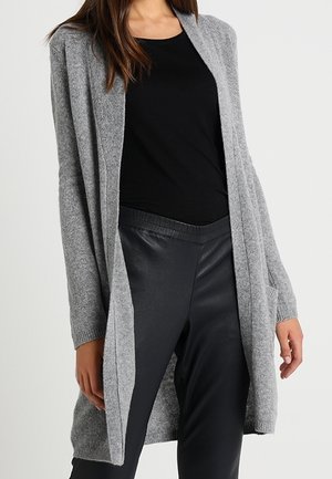 Kvinde iført en lang grå cardigan med åben front over en sort top og sorte læderbukser, stående mod en ensfarvet baggrund.