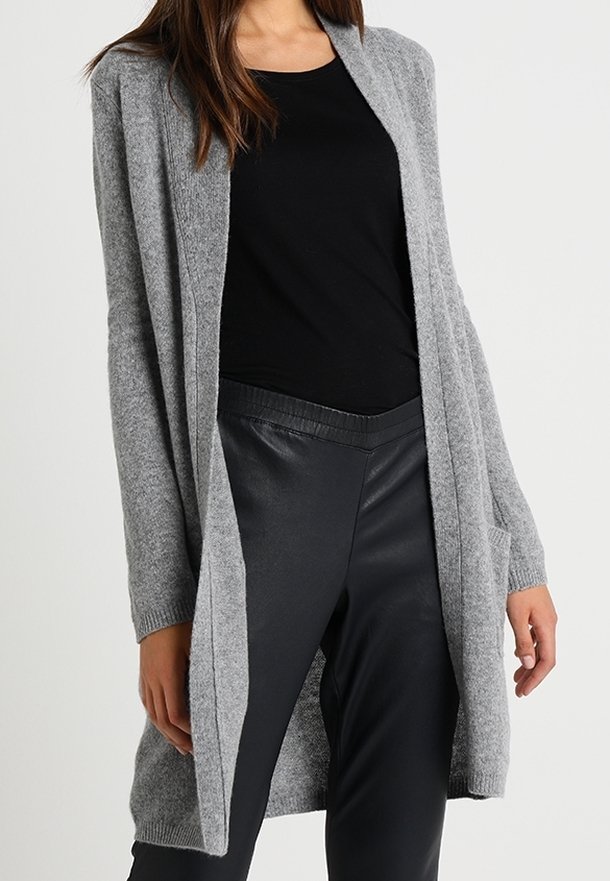 Femme portant un long cardigan gris ouvert sur le devant, par-dessus un haut noir et un pantalon en cuir noir, debout devant un fond uni.