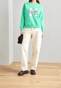 Mujer vistiendo un sudadera verde de Manhattan, pantalones de pierna ancha color crema, zapatos de tacón negros, sosteniendo un pequeño bolso negro, de pie en un suelo de madera.