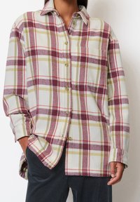 Flannel shirt in white, red, and olive green plaid pattern. Features a pointed collar, long sleeves, and button front with rounded hem.