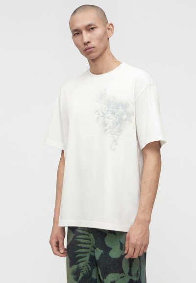 Άνδρας με ξυρισμένο κεφάλι που φοράει λευκό φαρδύ μπλουζάκι με διακριτικό floral μοτίβο και πράσινο παντελόνι με καμουφλάζ σχέδιο σε ανοιχτό γκρι φόντο.