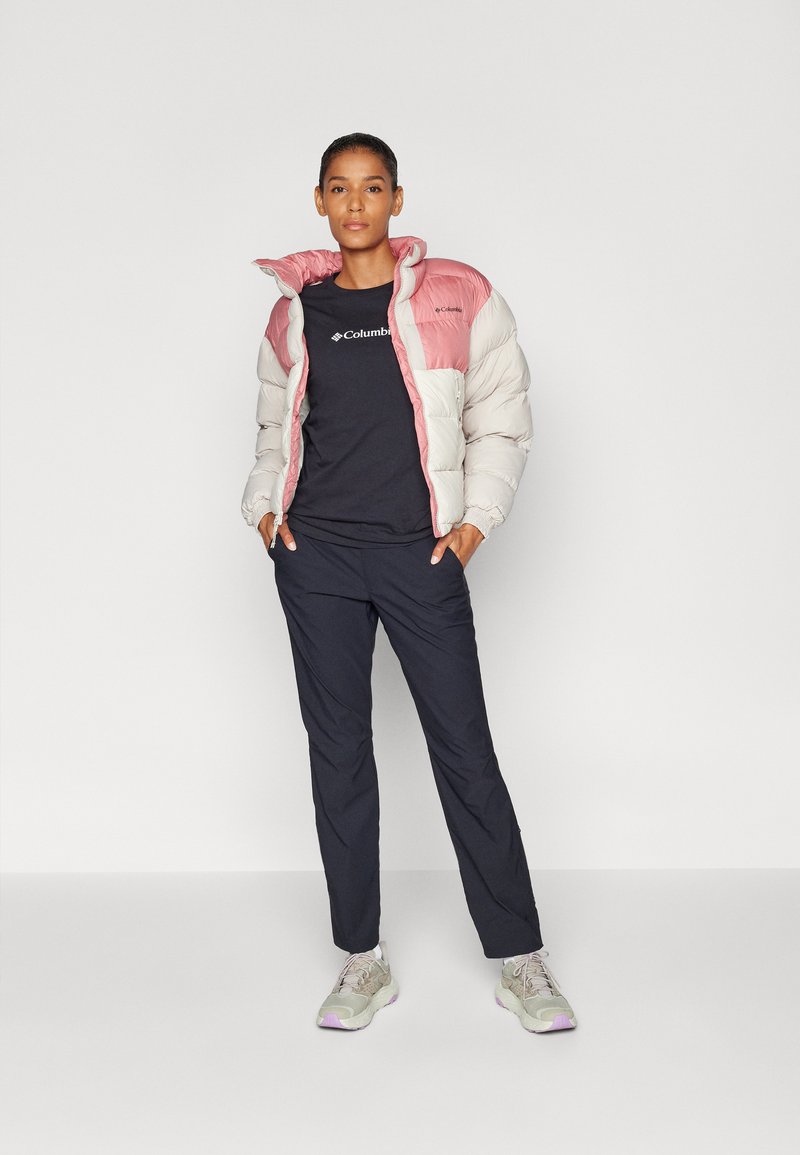 Chaqueta acolchada en gris claro con una sección superior rosa; camiseta negra con logo; pantalones oscuros; zapatillas gris claro con detalles.