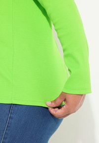 Langärmliges, geripptes Oberteil in leuchtendem Grün mit leicht geschwungenem Saum. Eine Hand hält den unteren Rand. Getragen mit blauen Jeans.