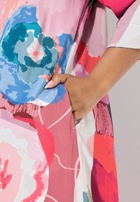 Kolorowa sukienka w kwiaty w odcieniach różu, czerwieni, niebieskiego i zieleni, z boczną kieszenią i miękką fakturą materiału. Ręka spoczywa na kieszeni.